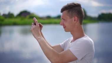 Side view portrait of happy Caucasian young man taking photos of summer river standing outdoors. Positive handsome guy photographing on smartphone in slow motion. Travelling and life balance