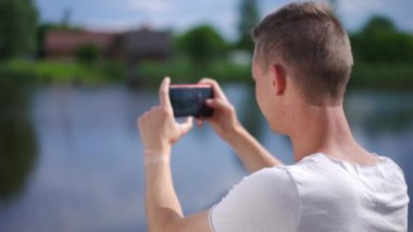 Shooting over shoulder of excited Caucasian man photographing nature standing on river bank outdoors. Happy young guy taking photos admiring water and green trees. Slow motion