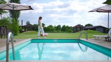 Side view confident gorgeous young Caucasian woman walking along swimming pool in slow motion sitting down on deck chair on the right. Wide shot positive satisfied tourist outdoors at resort