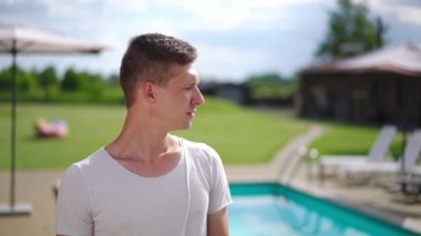 Medium shot satisfied relaxed young man standing at poolside enjoying sunny summer day and looking at camera smiling crossing hands. Portrait of Caucasian tourist posing in slow motion at resort