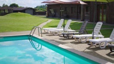 Blue swimming pool water with reflection of summer sky and clouds outdoors. Row of deck chairs sunbeds on poolside at tourist resort on sunny day with no people. Vacations concept