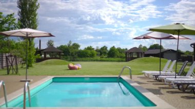 Wide shot blue swimming pool with green meadow and white clouds on sky at background. Cozy neat tourist resort in sunshine outdoors on summer day. No people