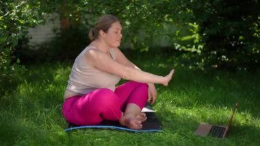 Side view beautiful plus-size sportswoman waving talking at laptop video chat smiling sitting on exercise mat in summer garden. Happy motivated Caucasian overweight woman training online outdoors
