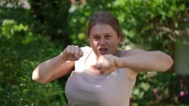 Front view portrait of angry plus-size woman looking at camera boxing shadow in slow motion. Motivated Caucasian overweight sportswoman training working out in summer garden outdoors posing