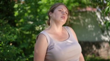 Tired overweight sportswoman hanging towel on shoulder wiping sweat off forehead in slow motion. Portrait of exhausted motivated Caucasian plus-size woman training in garden. Self improvement concept