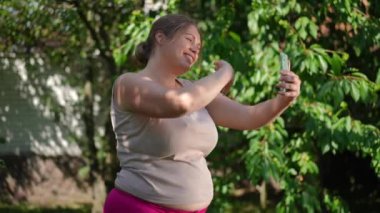 Side view joyful plus-size woman waving at smartphone video chat talking in slow motion. Smiling positive motivated Caucasian overweight sportswoman blogging in summer garden. Inspiration and sport