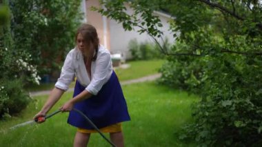 Overweight Caucasian woman watering green lawn with water hose in slow motion. Portrait of plus-size gardener pouring water on meadow in backyard garden on summer day. Horticulture and gardening