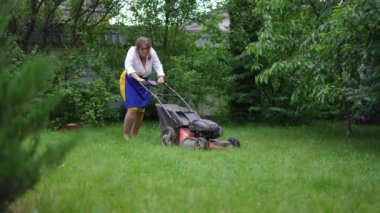Motivated plus-size woman trimming grass in garden with lawn mower walking in slow motion pushing equipment. Wide shot portrait of Caucasian overweight gardener working on summer day outdoors