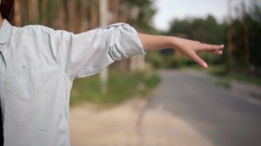 Unrecognizable young Caucasian woman gesturing hitchhiking on suburban road in forest. Slim female hand stretched in gesture catching car outdoors. Adventures and travelling concept