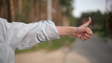 Close-up female hand gesturing thumb up hitchhiking with blurred cars passing at background. Unrecognizable young confident Caucasian woman on suburban road. Trips and travelling