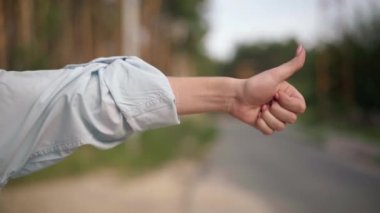 Close-up hand of unrecognizable woman hitchhiking gesturing thumb up on empty road at forest leaving. Unsatisfied stressed young Caucasian tourist outdoors alone