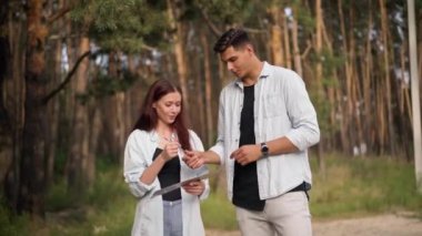 Loving positive young couple talking smiling writing destination on cardboard standing at forest. Front view Caucasian hitchhikers enjoying trip outdoors on vacations. Journey and enjoyment concept