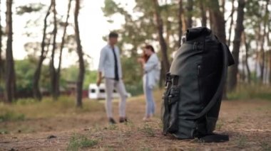 Close-up black rucksack in forest with blurred happy couple talking standing at background. Unrecognizable travelers discussing destination enjoying trip outdoors. Hiking and eco-tourism concept