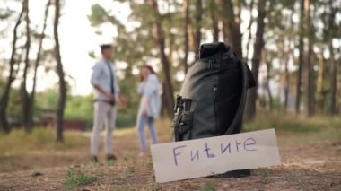 Close-up Future banner and backpack with blurred cheerful Caucasian couple laughing talking standing at background. Happy relaxed positive man and woman enjoying eco-tourism in forest. Lifestyle joy