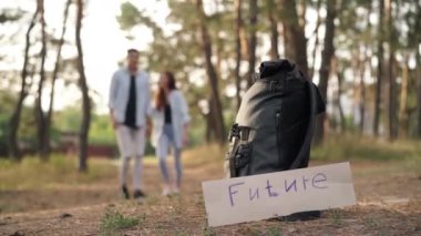 Future banner at backpack in autumn forest and blurred couple passing leaving at background. Carefree happy Caucasian boyfriend and girlfriend enjoying vacations outdoors. Relationship and leisure
