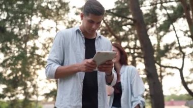 Focused handsome boyfriend searching route direction online as girlfriend talking on phone smiling. Happy positive Caucasian couple of hikers travelling in forest outdoors