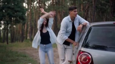 Stressed handsome man leaning on broken car with woman jumping gesturing calling help. Front view of Caucasian boyfriend and girlfriend with automobile in forest. Trip problem