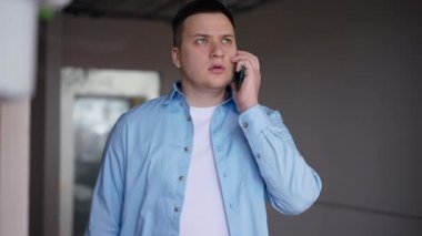 Satisfied young man picking up phone talking smiling standing in new flat. Portrait of Caucasian real estate owner indoors in new apartment. Renovation and moving concept