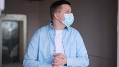 Medium shot portrait of thoughtful young man in coronavirus face mask standing in apartment under construction looking around. Caucasian new flat owner planning repair indoors on Covid-19 pandemic