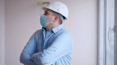 Professional young foreman in Covid-19 face mask crossing hands looking around standing in building under construction. Thoughtful expert Caucasian man in hard hat planning repair in new flat