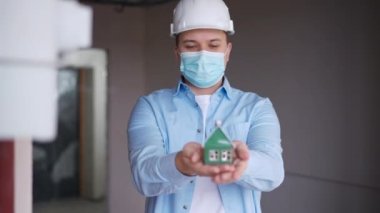Builder in coronavirus face mask and hard hat posing with toy house in new flat indoors. Front view portrait of young positive Caucasian man looking at camera smiling. Construction and expertise