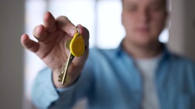 Close-up apartment key in male Caucasian hand with blurred satisfied owner at background. Unrecognizable confident young man posing indoors in new flat. Real estate purchase concept