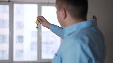 Blurred young man stretching key looking out the window in new apartment. Shooting over shoulder of Caucasian flat owner indoors. Ownership and real estate purchase concept