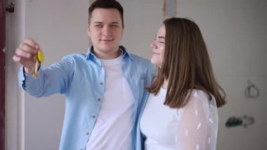 Loving young Caucasian couple standing in new flat under construction stretching apartment key looking away. Happy beautiful woman and handsome man moving in planning repair