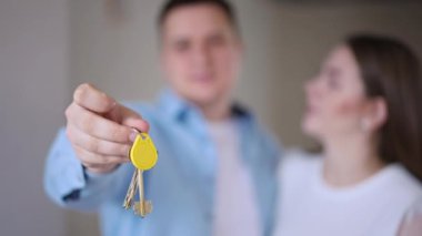 Close-up new flat key with blurred happy young couple talking at background smiling. Satisfied Caucasian man and woman bragging apartment ownership standing indoors