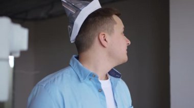 Positive man in paper hat looking around smiling standing in new flat indoors. Portrait of Caucasian young guy planning repair thinking. Renovation and ownership concept
