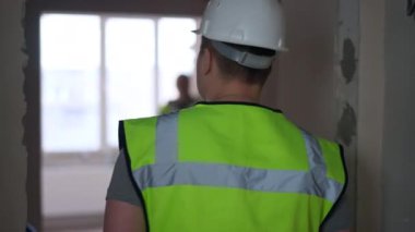 Tracking shot of Caucasian man in green vest walking to positive woman standing in new apartment. Live camera follows confident architect talking with worker indoors in flat under construction