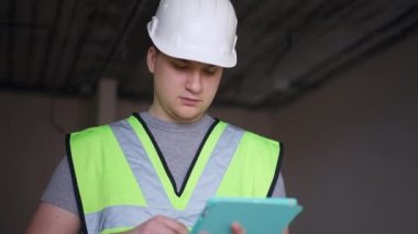 Bottom angle view of intelligent concentrated architect in hard hat using digital tablet looking around standing in new apartment indoors. Focused handsome Caucasian young man checking quality