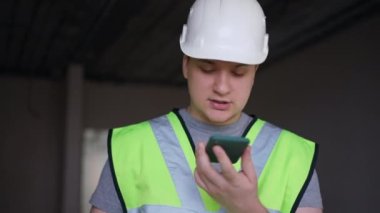 Portrait of dissatisfied Caucasian foreman talking at smartphone loudspeaker with annoyed facial expression. Young man in hard hat arguing standing indoors in new flat under construction