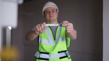 Measuring roulette tape in male hands with blurred man smiling at background looking at camera. Happy confident professional Caucasian builder posing in new flat indoors with equipment