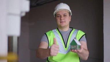 Medium shot front view of confident builder gesturing thumb up showing toy house standing indoors. Handsome young Caucasian man in hard hat and green vest posing in new apartment looking at camera