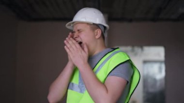 Overworking exhausted young builder yawning shaking hand leaving new apartment under construction. Portrait of tired Caucasian man walking away. Building and exhaustion