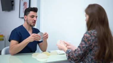 Middle Eastern doctor and Caucasian patient choosing breast implants talking sitting indoors. Confident expert man and curios woman discussing aesthetic surgery planning operation on augmentation