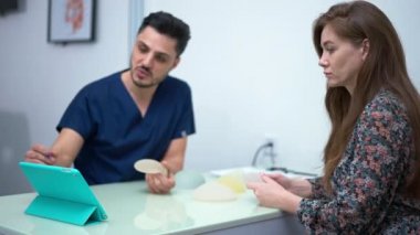 Side view focused Caucasian woman choosing breast implant sitting on the right with blurred Middle Eastern surgeon talking pointing at digital tablet. Concentrated patient consulting doctor indoors
