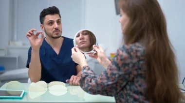 Thoughtful Caucasian woman reflecting in hand mirror planning aesthetic surgery with blurred Middle Eastern doctor talking at background smiling. Patient and surgeon on visit in medical clinic