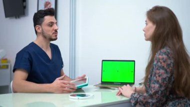 Professional doctor with digital tablet and green screen laptop talking with patient gesturing. Portrait of handsome Middle Eastern plastic surgeon consulting Caucasian woman in medical clinic