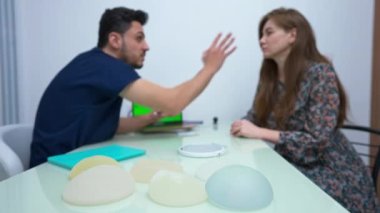 Close-up implants for breast augmentation in medical clinic with blurred doctor gesturing consulting patient at background. Confident Middle Eastern man explaining procedure to Caucasian woman