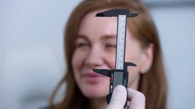 Close-up of vernier caliper in doctor hand with blurred Caucasian woman smiling at background. Brunette positive cheerful female patient consulting professional plastic surgeon for facelift