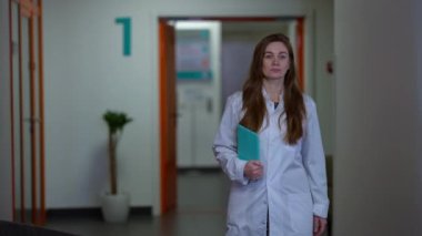 Adult Caucasian woman in white hospital gown walking in hallway stops looking around. Troubled female doctor with digital tablet thinking standing in corridor