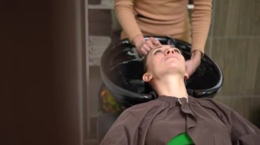 Front view slim young Caucasian woman sitting in hair salon with head in sink and unrecognizable hairdresser washing hair. Positive confident client and professional hairstylist in beauty parlor