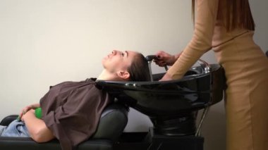 Side view slim young woman sitting at washing sink in hair parlor with unrecognizable hairstylist washing off shampoo in slow motion with water. Caucasian client and hairdresser in beauty salon