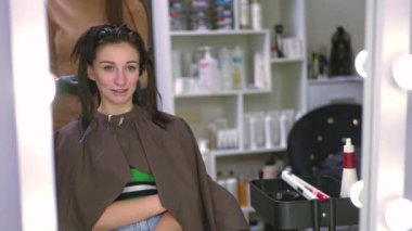 Positive woman reflecting in beauty parlor mirror smiling as professional hairstylist using infrared flat iron for keratin treatment. Charming Caucasian client and expert hairdresser in hair salon