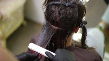 High angle view brunette long hair straightening with infrared flat iron. Unrecognizable Caucasian professional hairstylist applying keratin treatment on strand of woman in hair salon
