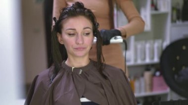 Reflection in mirror of focused woman talking as hairdresser using flat iron straightening hair. Front view of Caucasian slim young lady doing hairstyle in beauty salon indoors chatting