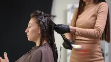 Side view young woman talking with hairdresser straightening hair with flat iron in beauty salon. Caucasian female client and hairstylist doing hairdo with infrared appliance in hair parlor