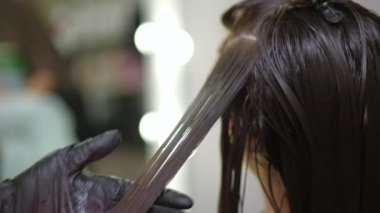 Close-up brunette hair strand with hand in gloves using infrared flat iron. Unrecognizable professional expert hairstylist doing keratin treatment for young Caucasian lady passing closing camera